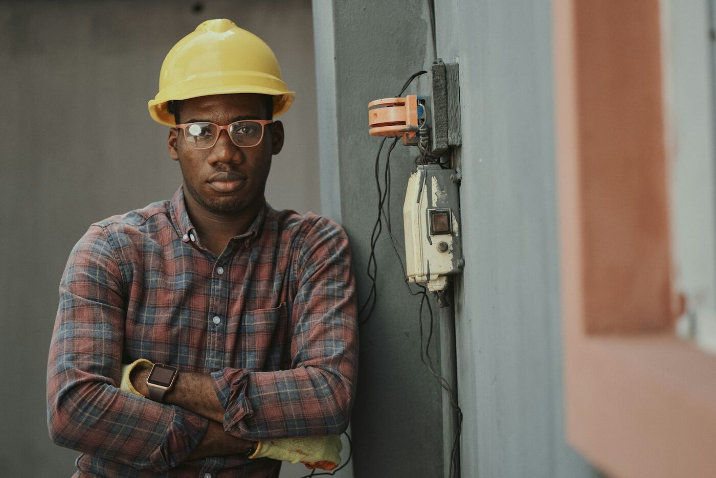 An electrician in a yellow hard hat, arms crossed, in front of a wall of meters