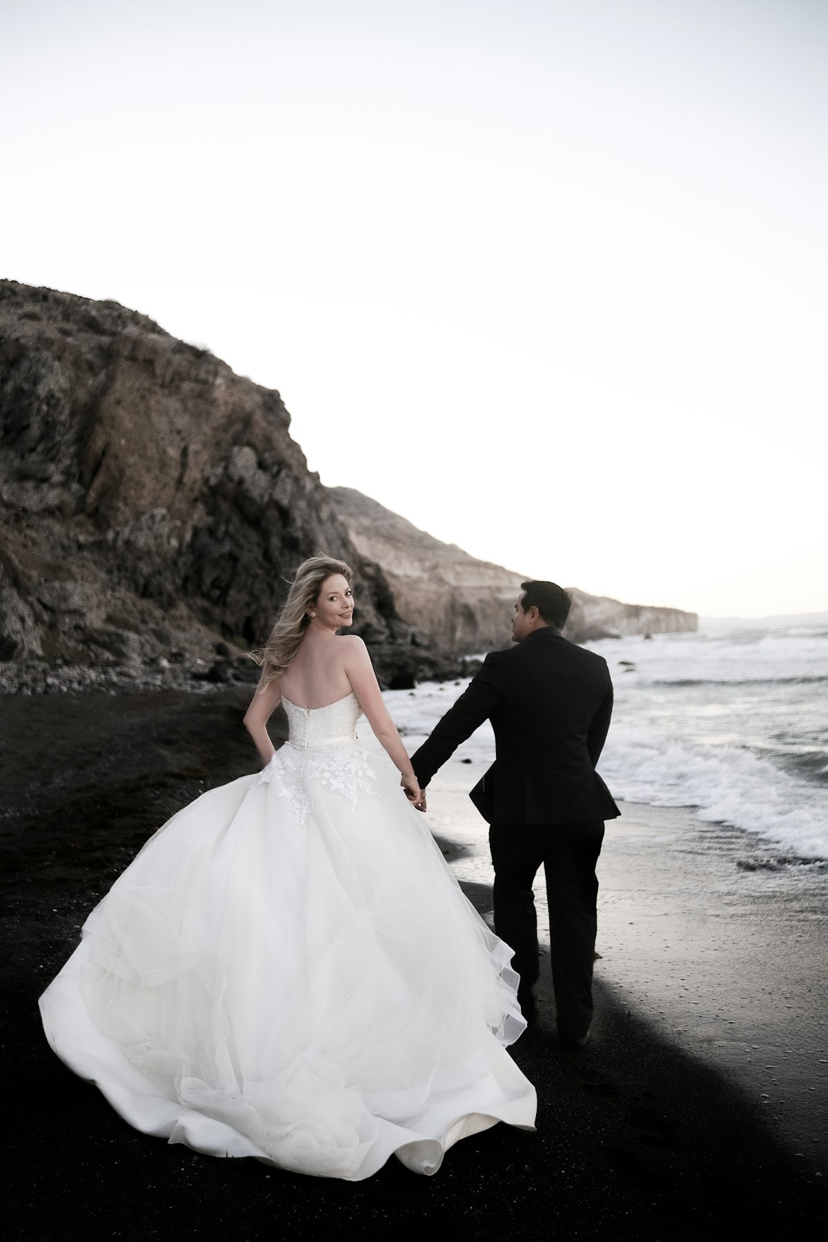 A bride and groom walking hand in hand along a black sand beach
